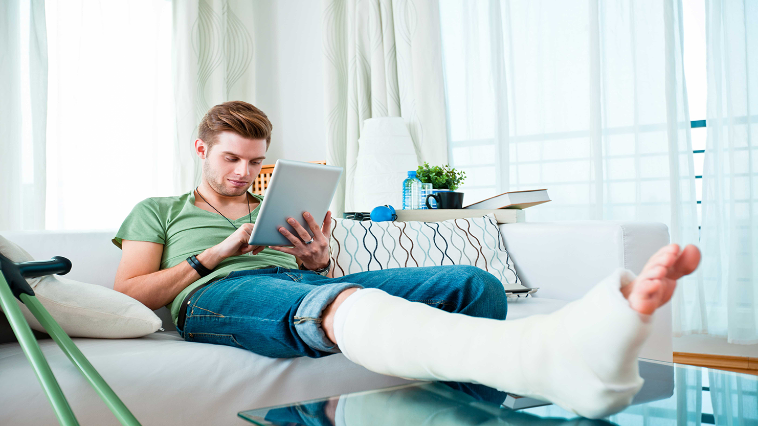 Man with a leg cast, sitting on a couch, looking at a tablet