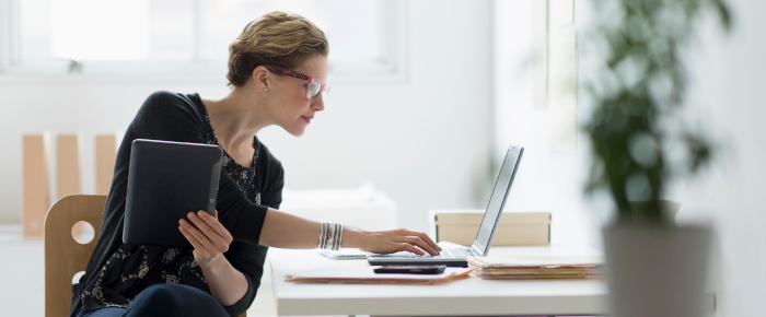 Woman sitting at a desk, holding a tablet and typing on a laptop