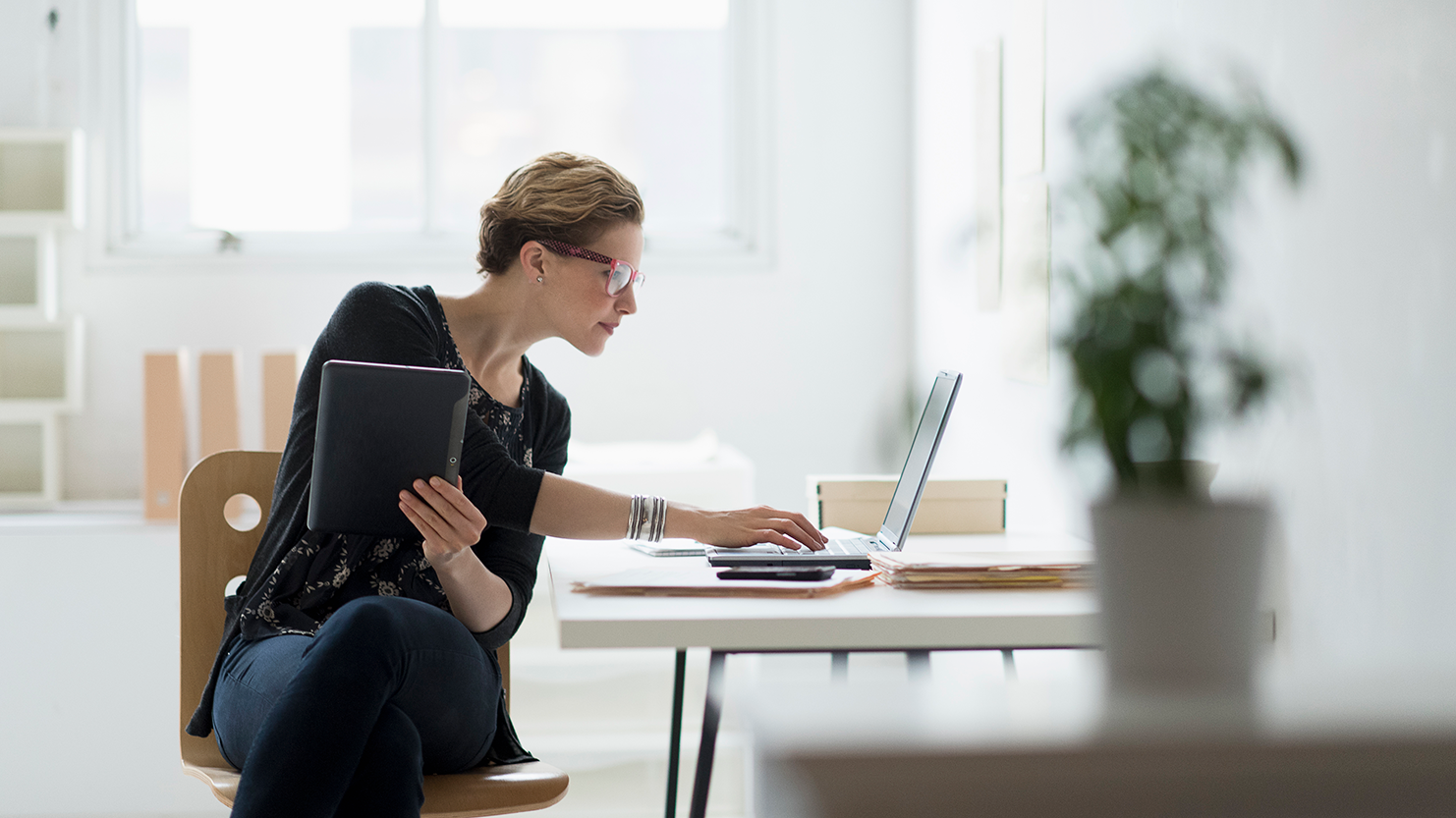 Woman sitting at a desk, holding a tablet and typing on a laptop